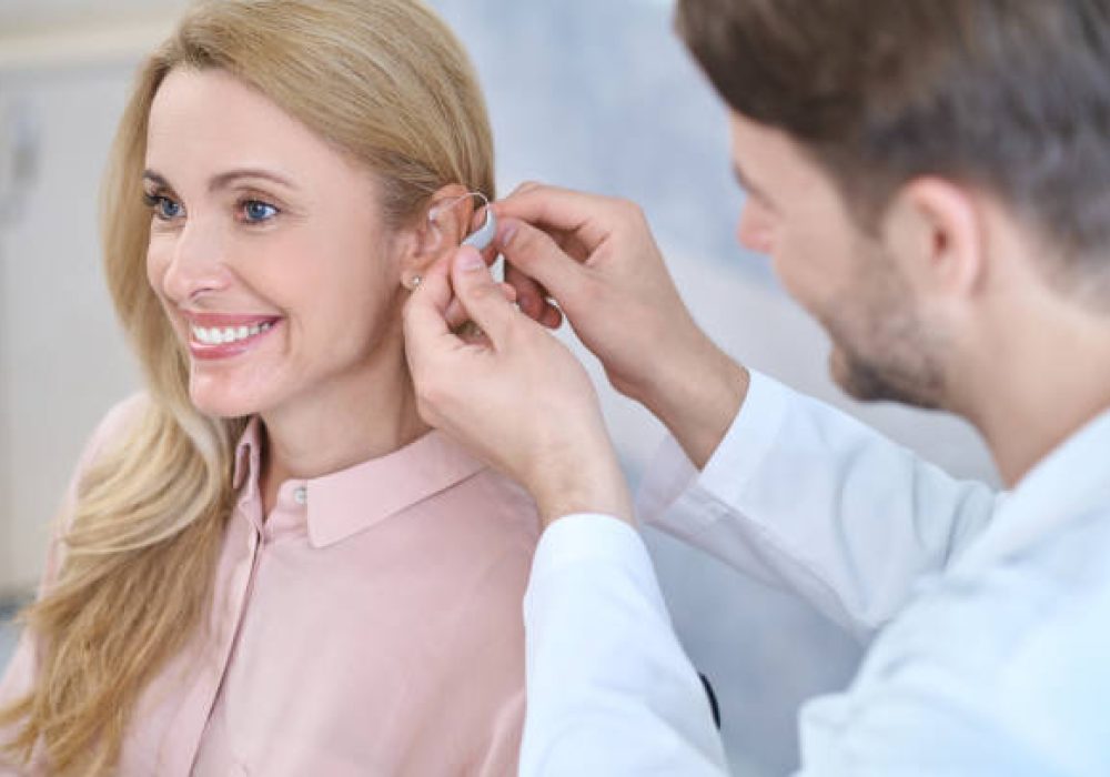 IMG_5151 Fitting. Man in a white coat putting hearing aid on ear of smiling middle-aged woman with long blond hair sitting in medical office